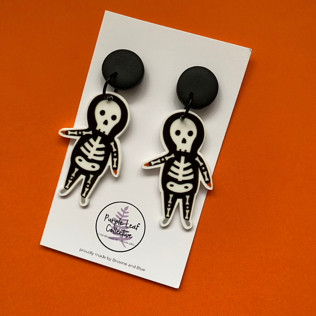 Skeleton-themed earrings on a card with 'Purple Leaf Collective' branding against an orange background.