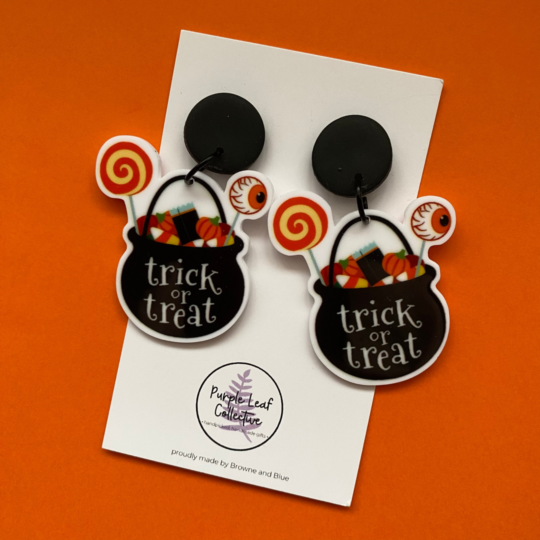 Halloween-themed earrings with 'trick or treat' text on a branded card against an orange background.