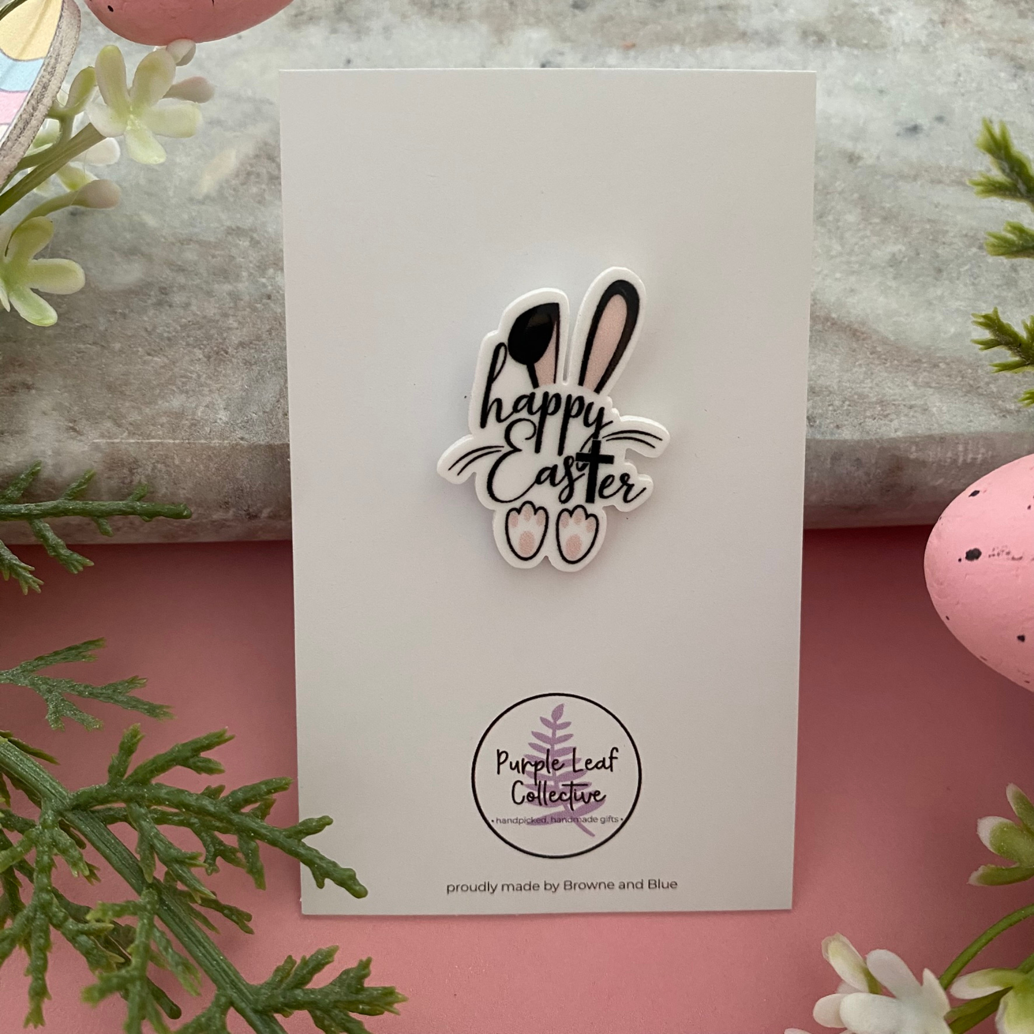 A handmade brooch featuring the words Happy Easter decorated with white rabbit ears on top and cute bunny feet at the bottom.