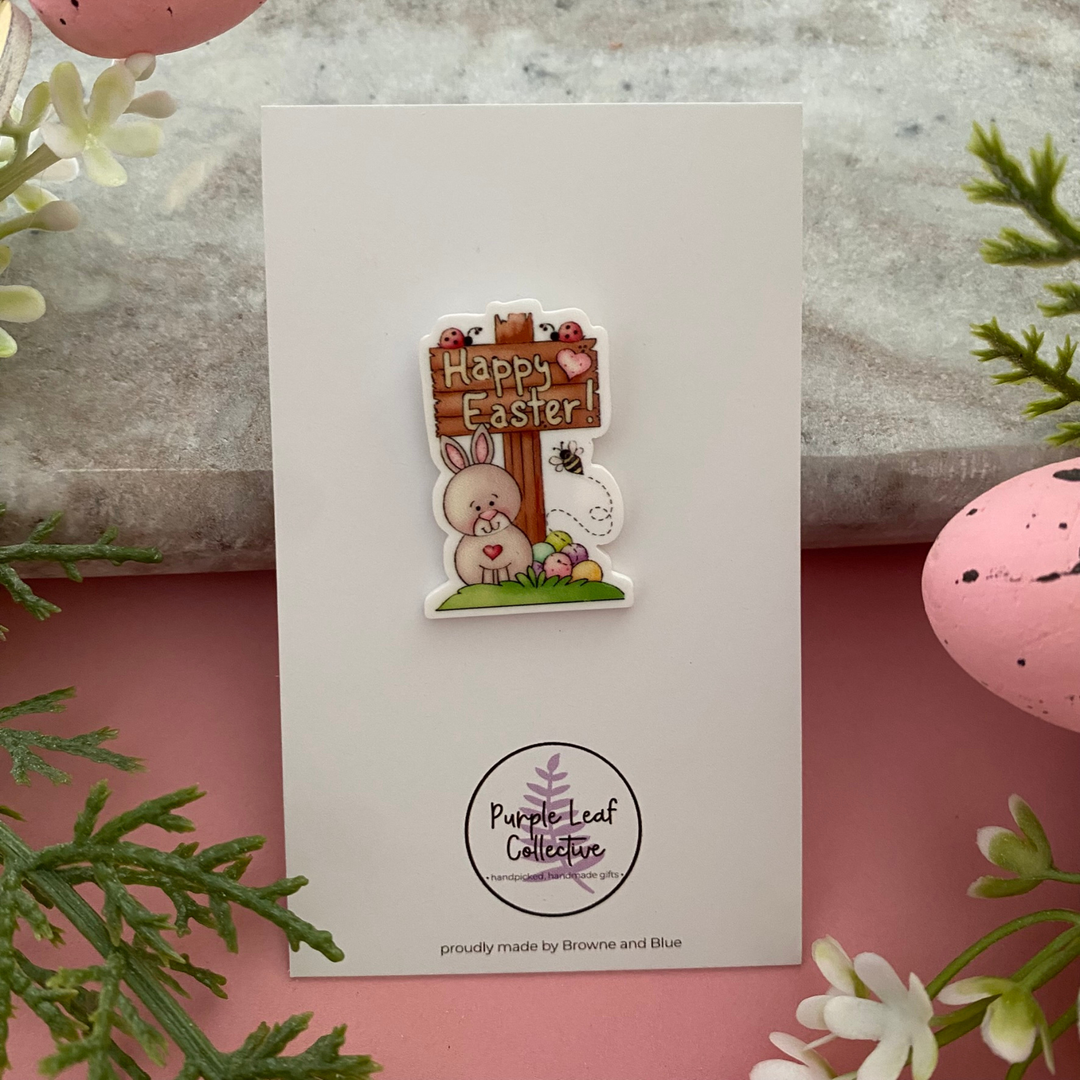 A detailed brooch featuring a white rabbit, colourful Easter eggs, a bee and a ladybug next to a wooden-style sign that reads Happy Easter.