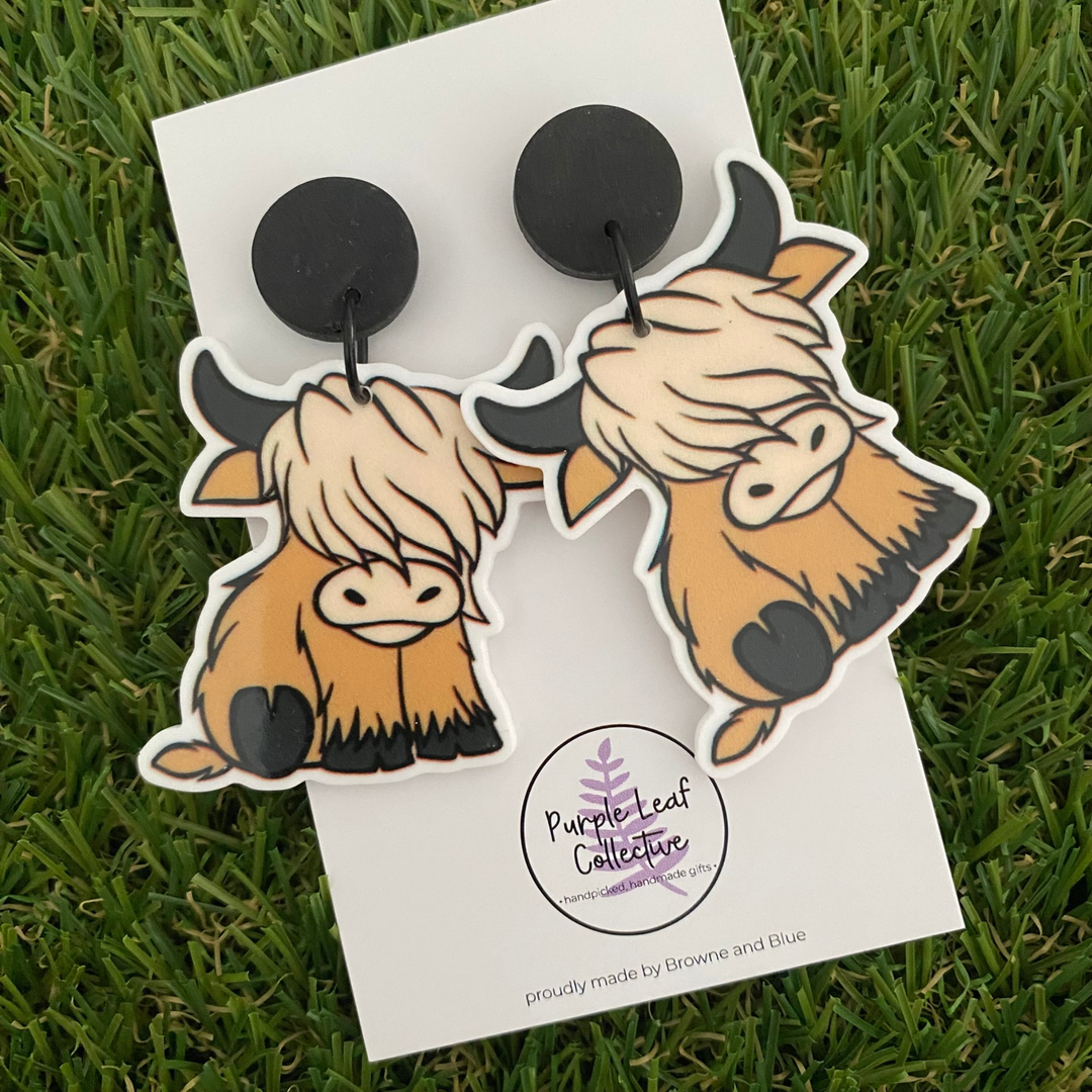 A pair of blonde haired highland cow dangle earrings displayed on a backing card.