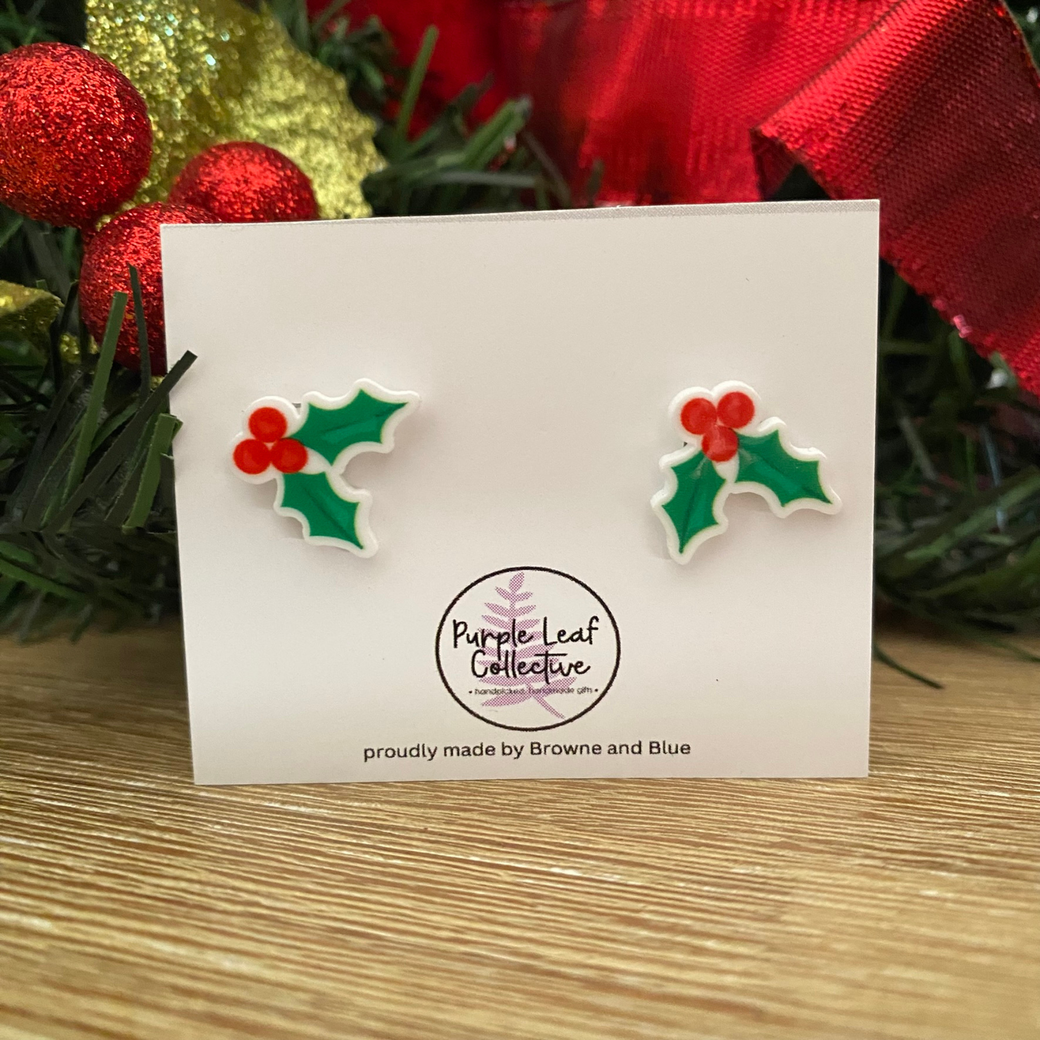 Holly leaf earrings on a card with Christmas decorations in the background.