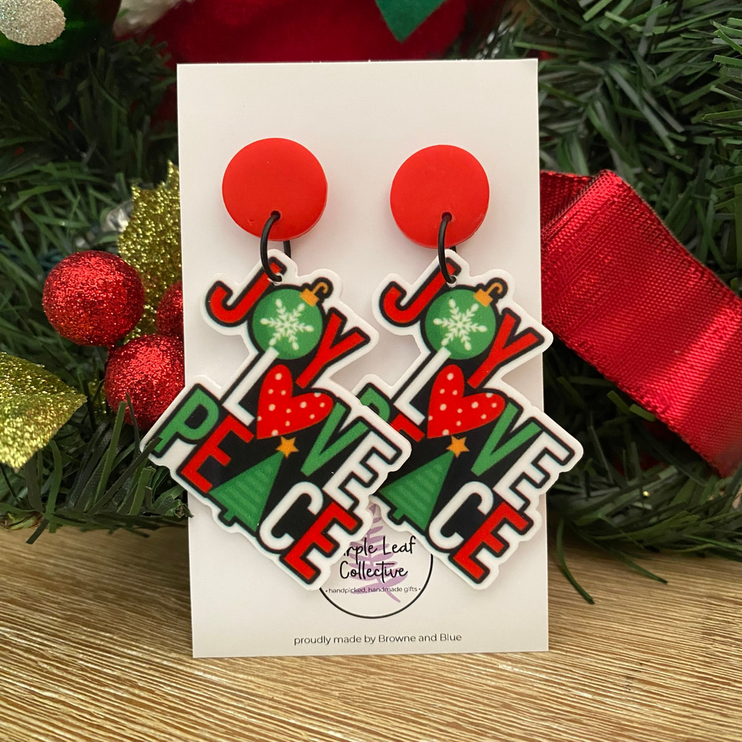 Pair of earrings with 'Joy Peace Love' text on a card with Christmas decorations in the background.