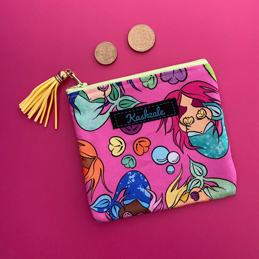 A small coin purse perfect for coins and small essentials featuring colourful mermaids by Kasey Rainbow on hot pink fabric and a tassel on the zipper.