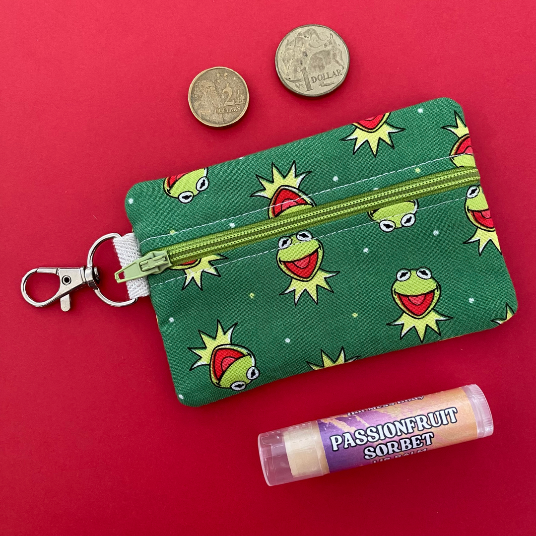 A small clip coin purse perfect for coins and small essentials featuring playful images of Kermit and his iconic green grin set on green fabric.