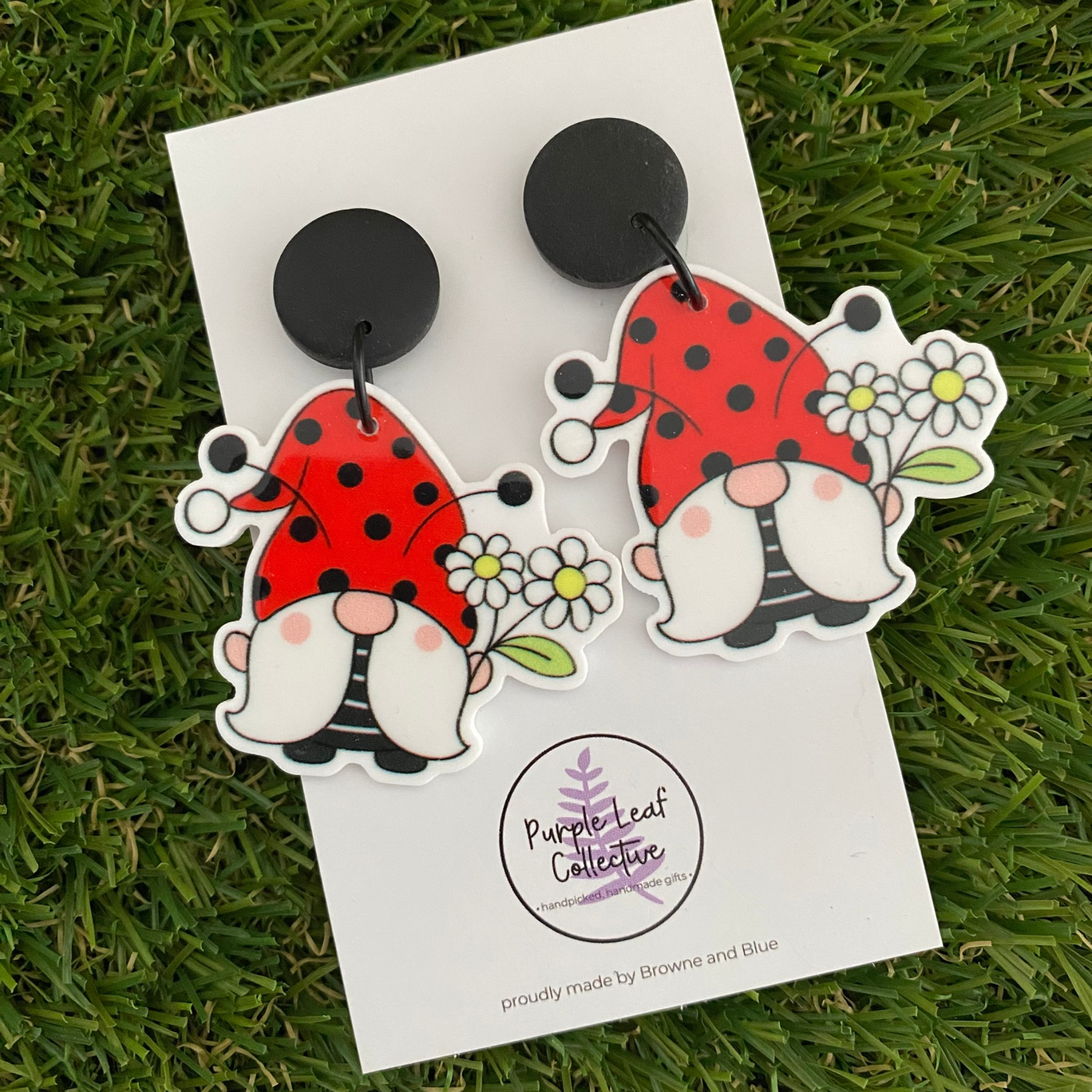 Pair of dangle earrings featuring two gnomes wearing ladybug-inspired hats and holding a bunch of flowers, set on sleek black clay tops.