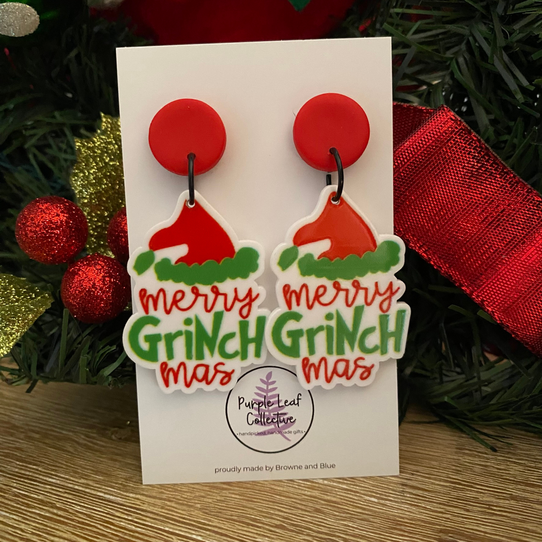 Pair of 'Merry Grinchmas' earrings on a card with Christmas decorations in the background.
