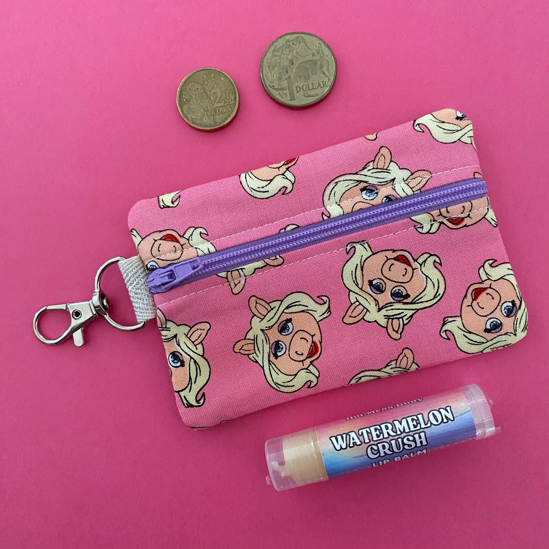 A small clip coin purse perfect for coins and small essentials adorned with playful images of Miss Piggy in all her fashionable glory on a pink fabric background.