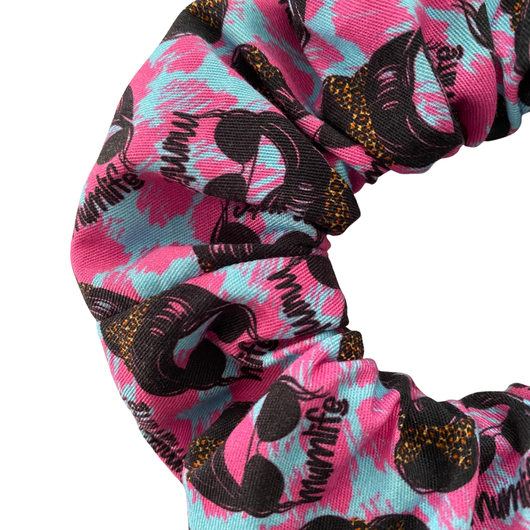Close up view of a hot pink and blue fabric scrunchie with images of a woman rocking sunglasses and a messy bun, complete with her own bow scrunchie.