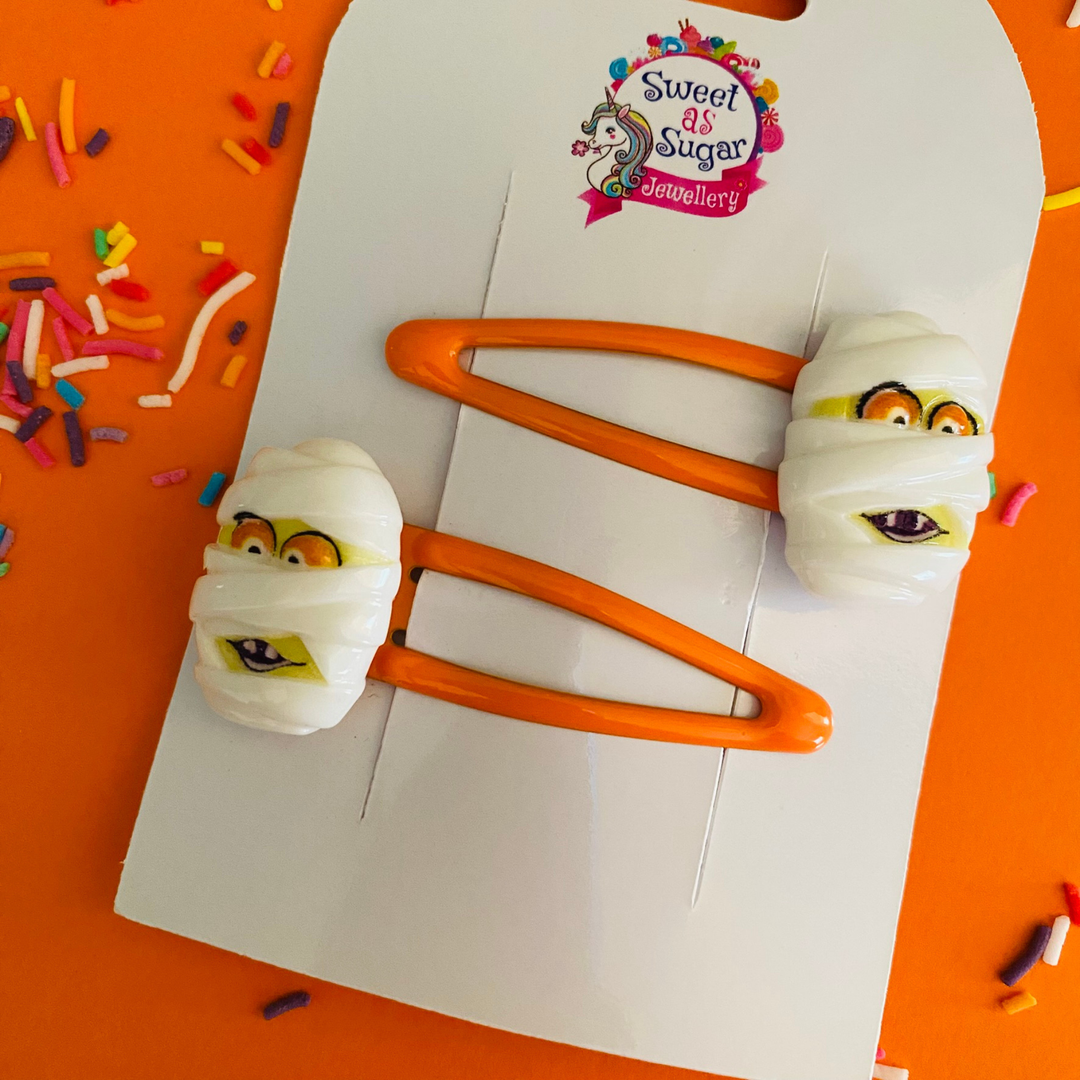 Mummy hair clips feature a classic bandage face—complete with eyes and teeth—set on a vibrant orange hair clip.