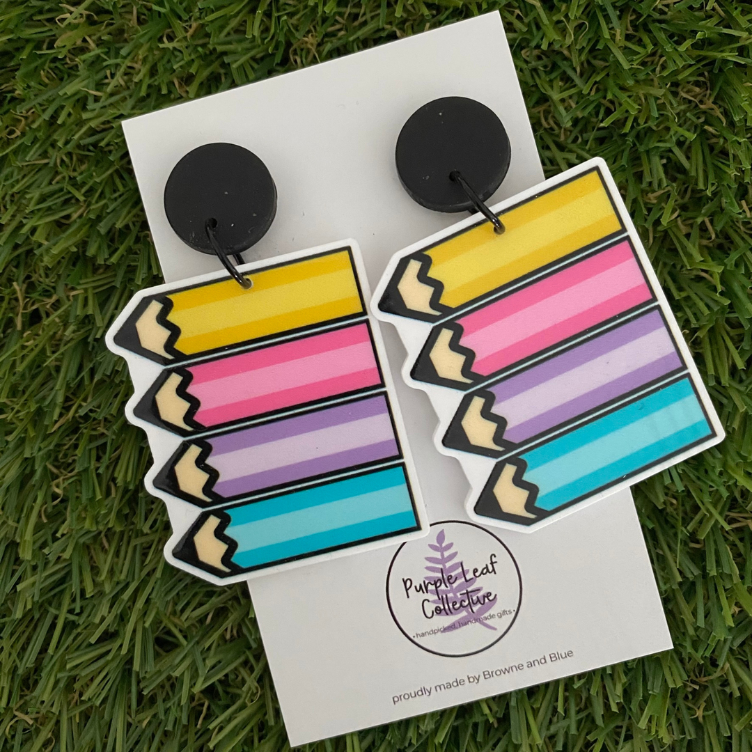 Dangle earrings featuring a cluster of four pencils in soft yellow, pink, purple, and blue tones set on sleek black clay tops.