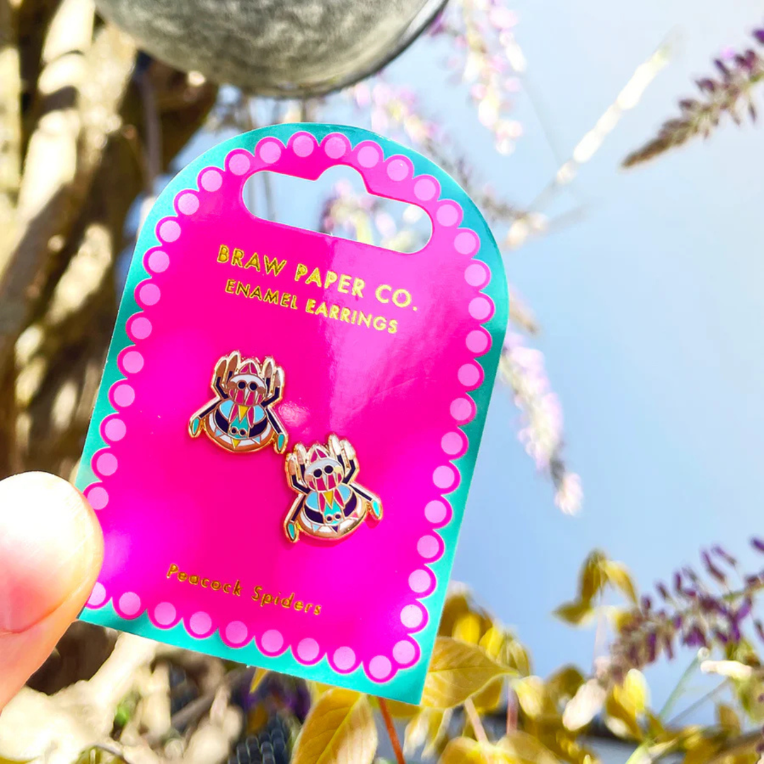 A pair of Peacock Spider enamel stud earrings on backing card on a flower background.