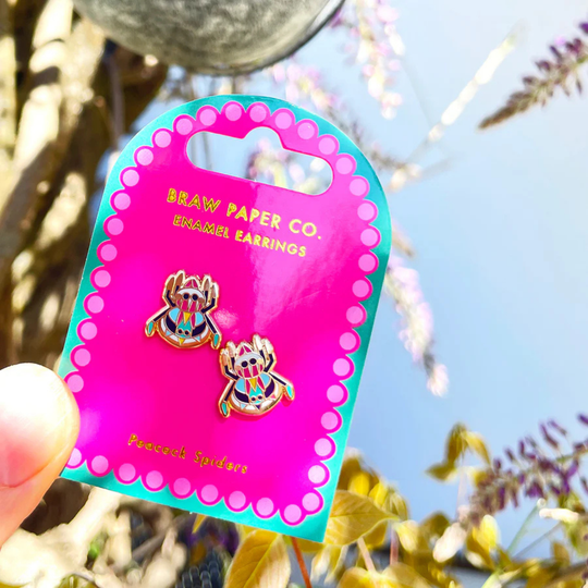 A pair of Peacock Spider enamel stud earrings on backing card on a flower background.