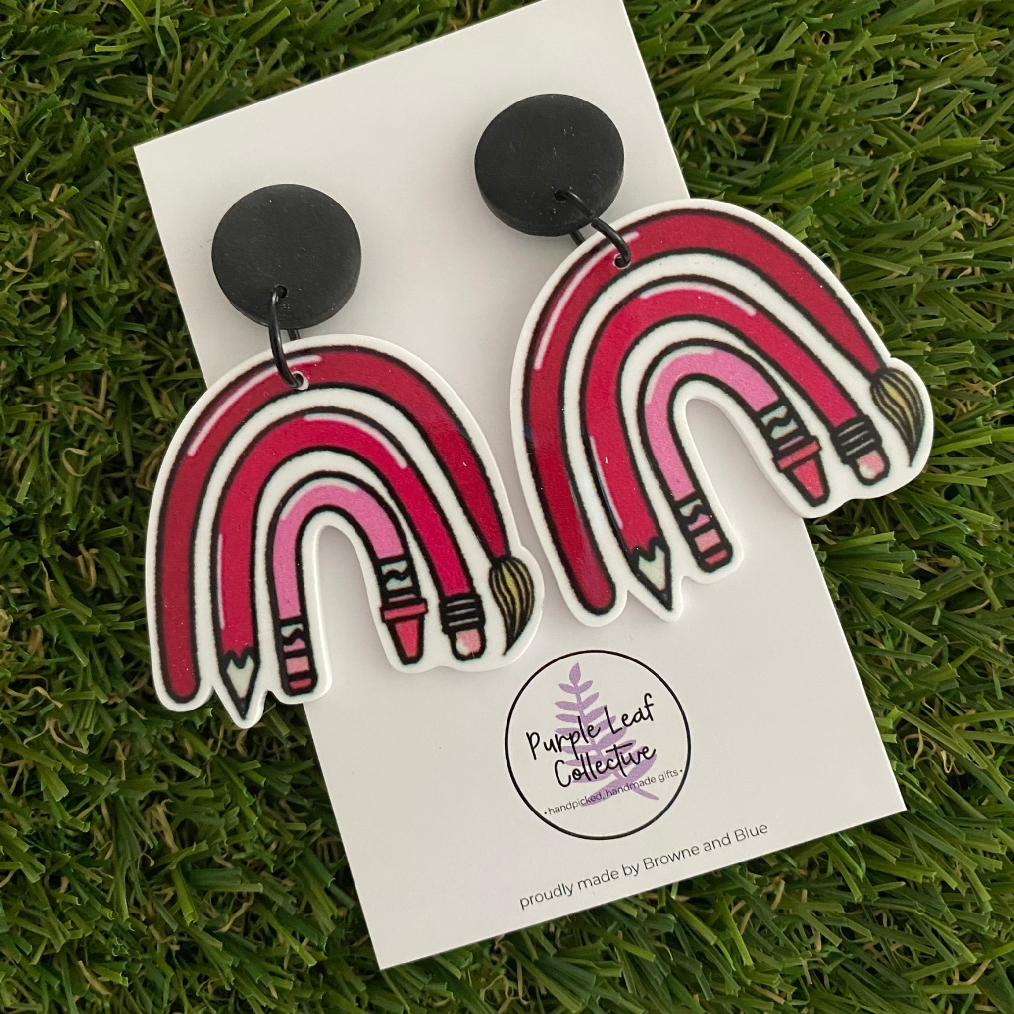 Pair of earrings featuring a paint brush, pencil, and crayon arranged in a pink rainbow pattern set on black clay tops.