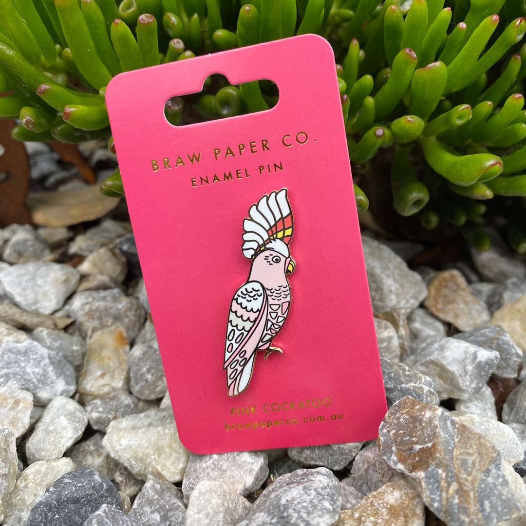 The Pink Cockatoo Enamel Pin featuring intricate details and showcasing a salmon pink face, throat, chest, and belly. Its standout feature is its stunning crest, adorned with bold red and yellow stripes.
