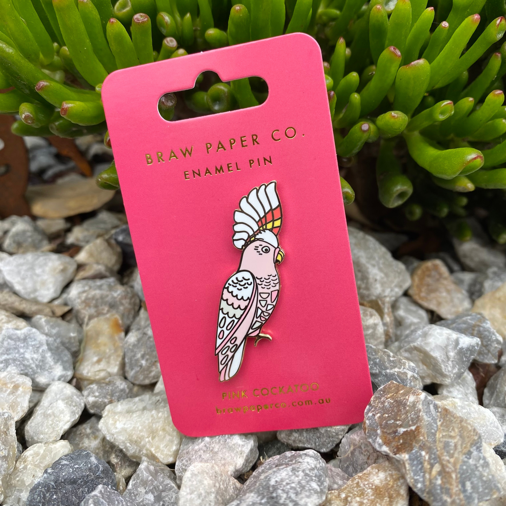 The Pink Cockatoo Enamel Pin featuring intricate details and showcasing a salmon pink face, throat, chest, and belly. Its standout feature is its stunning crest, adorned with bold red and yellow stripes.