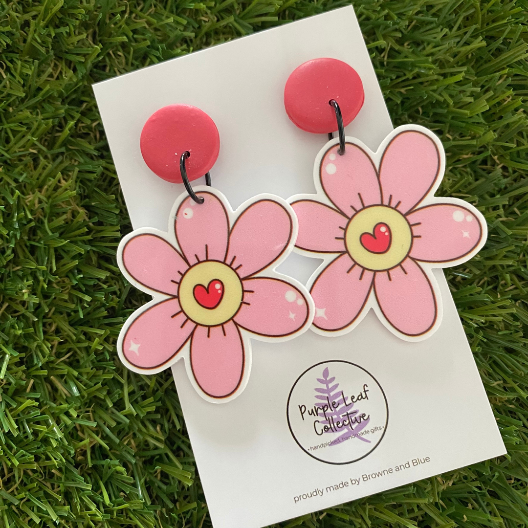 A pair of dangle earrings, featuring a pair of pink flowers with a red heart in the centre set on dark pink clay tops.