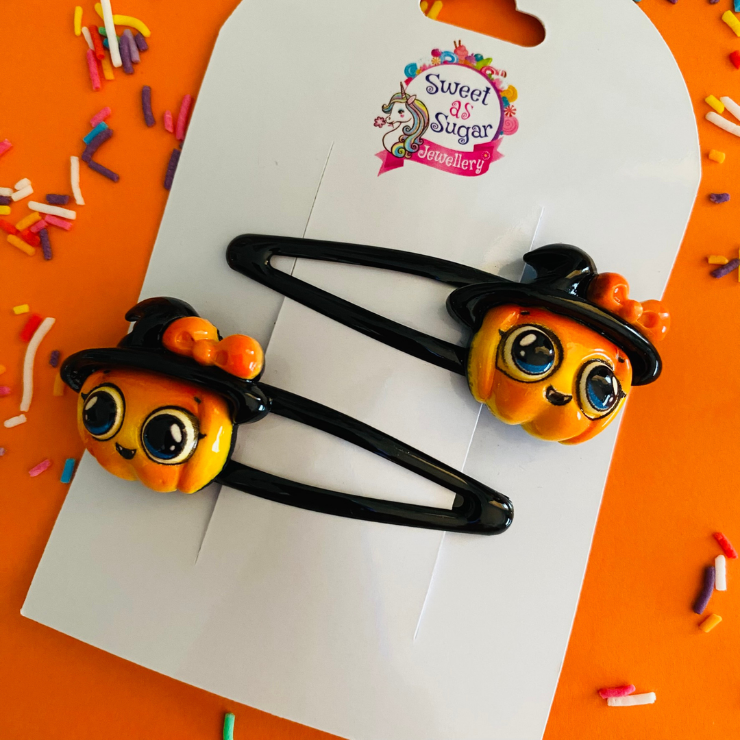 Pumpkin hair clips feature a classic jack-o'-lantern face adorned with a black witch’s hat and an orange bow, all set on a black hair clip.
