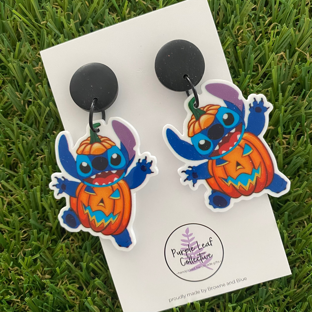 A pair of dangle earrings featuring the blue alien Stitch wearing a pumpkin suit and hat on black clay tops.