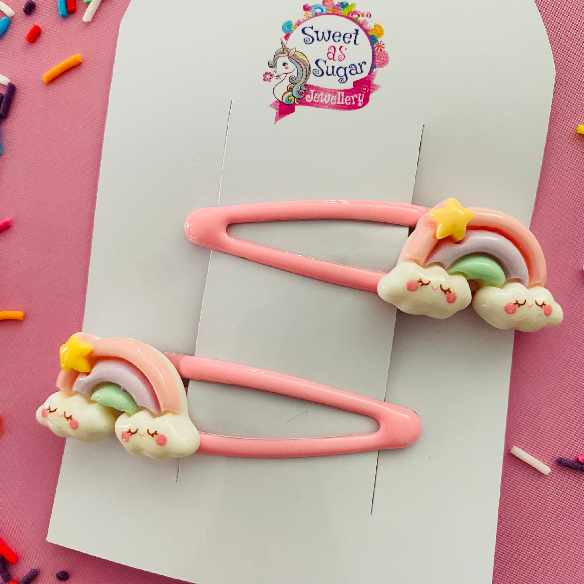 A pair of pink snap clips featuring  a pastel coloured rainbow, sitting on clouds with a yellow star.