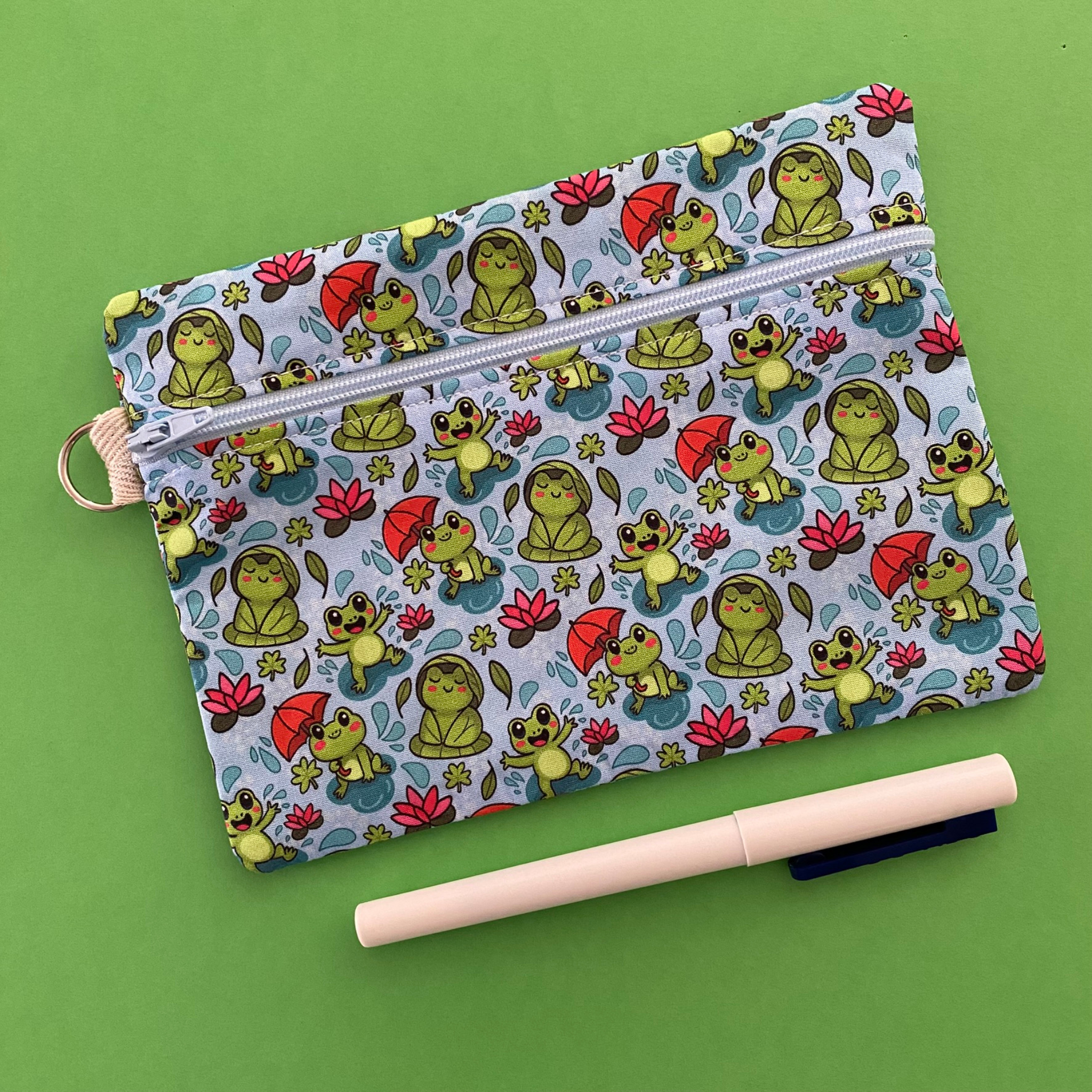 Zipper pouch featuring cute frogs in the rain holding umbrellas and sitting on lily pads on a crisp blue fabric.