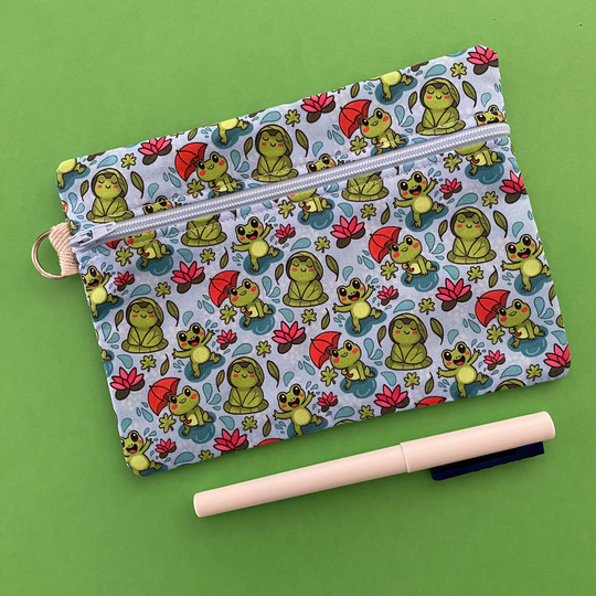 Zipper pouch featuring cute frogs in the rain holding umbrellas and sitting on lily pads on a crisp blue fabric.