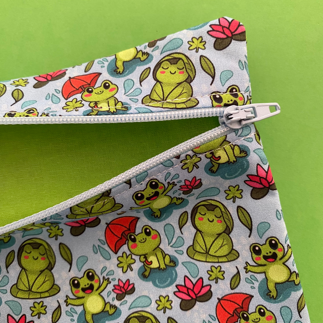 Zipper pouch featuring cute frogs in the rain holding umbrellas and sitting on lily pads on a crisp blue fabric featuring lime green lining.
