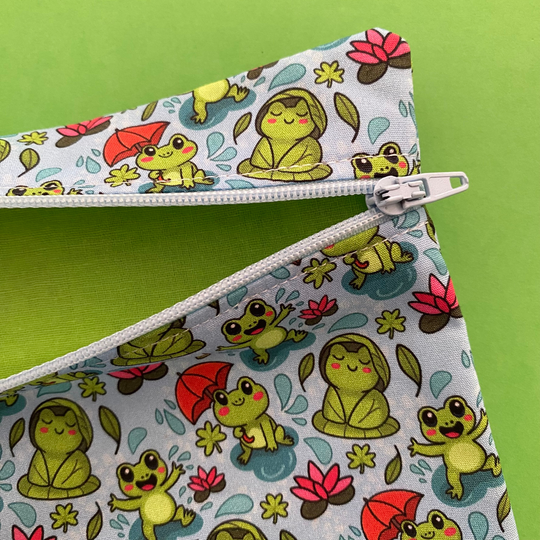 Zipper pouch featuring cute frogs in the rain holding umbrellas and sitting on lily pads on a crisp blue fabric featuring lime green lining.