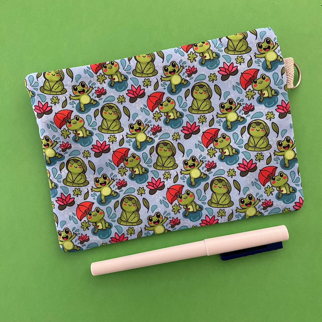 Reverse view of a zipper pouch featuring cute frogs in the rain holding umbrellas and sitting on lily pads on a crisp blue fabric.