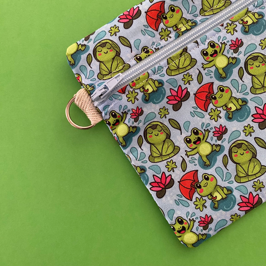 Side view of a zipper pouch featuring cute frogs in the rain holding umbrellas and sitting on lily pads on a crisp blue fabric.