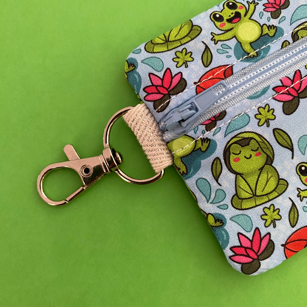 Clasp view of a mini pouch with images of frogs holding umbrellas and lily pads on a crisp blue fabric.
