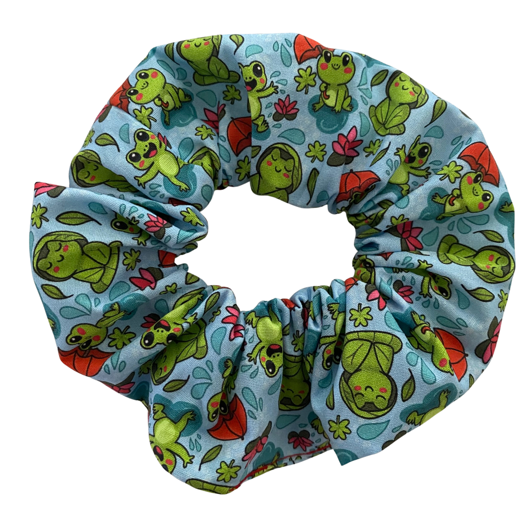 A blue fabric scrunchie with images of frogs in the rain holding umbrellas and sitting on lily pads.