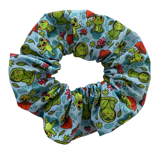 A blue fabric scrunchie with images of frogs in the rain holding umbrellas and sitting on lily pads.