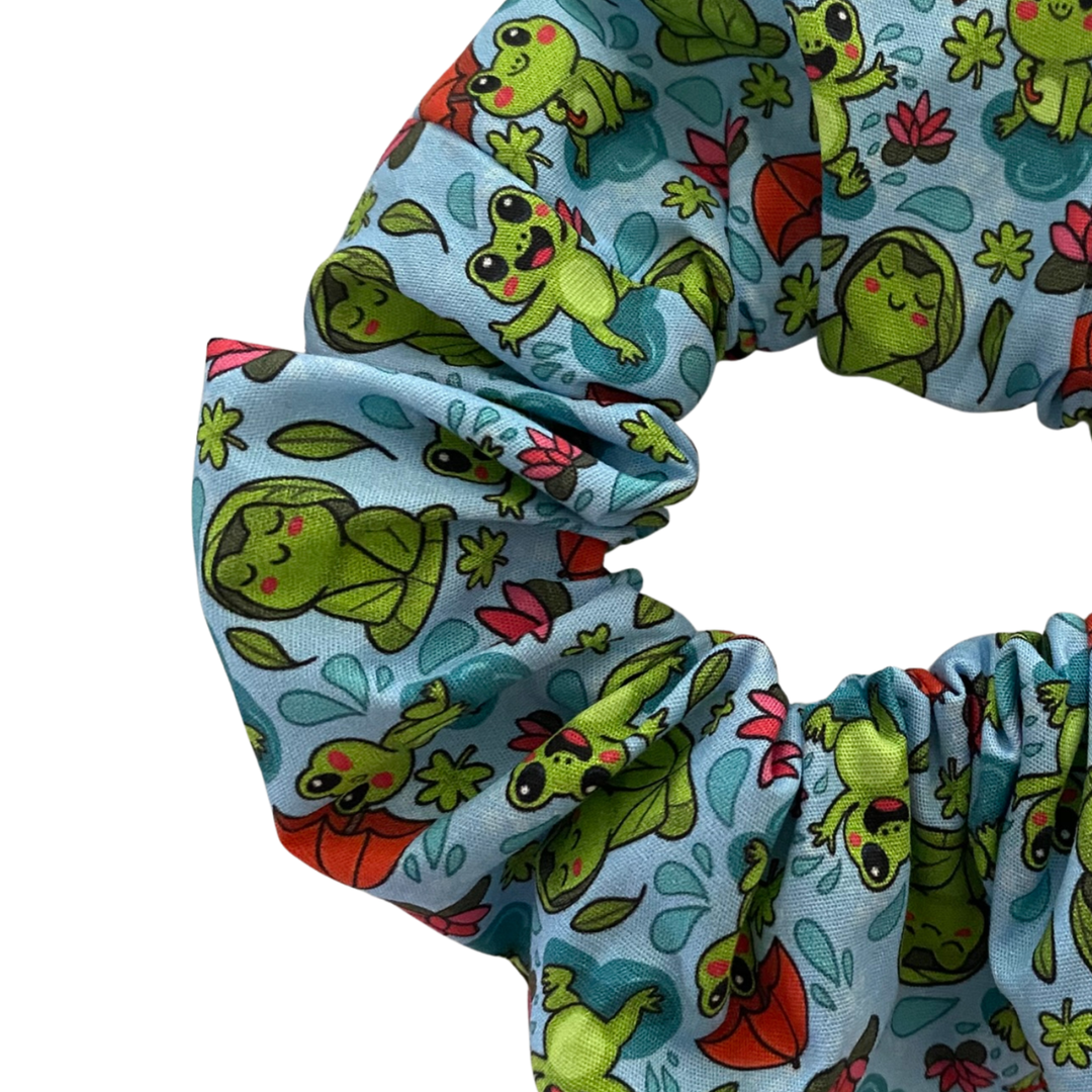 Close up view of a blue fabric scrunchie with images of frogs in the rain holding umbrellas and sitting on lily pads.