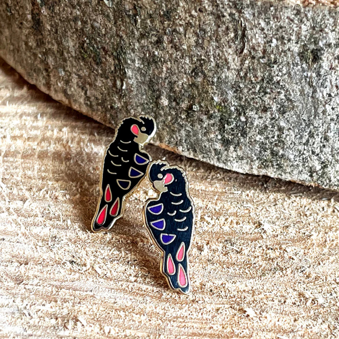 A pair of Red-tailed Black Cockatoo enamel stud earrings on a wooden surface with a stone background.