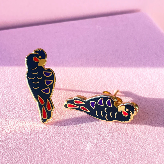 A pair of Red-tailed Black Cockatoo enamel stud earrings on a pink surface with shadows.