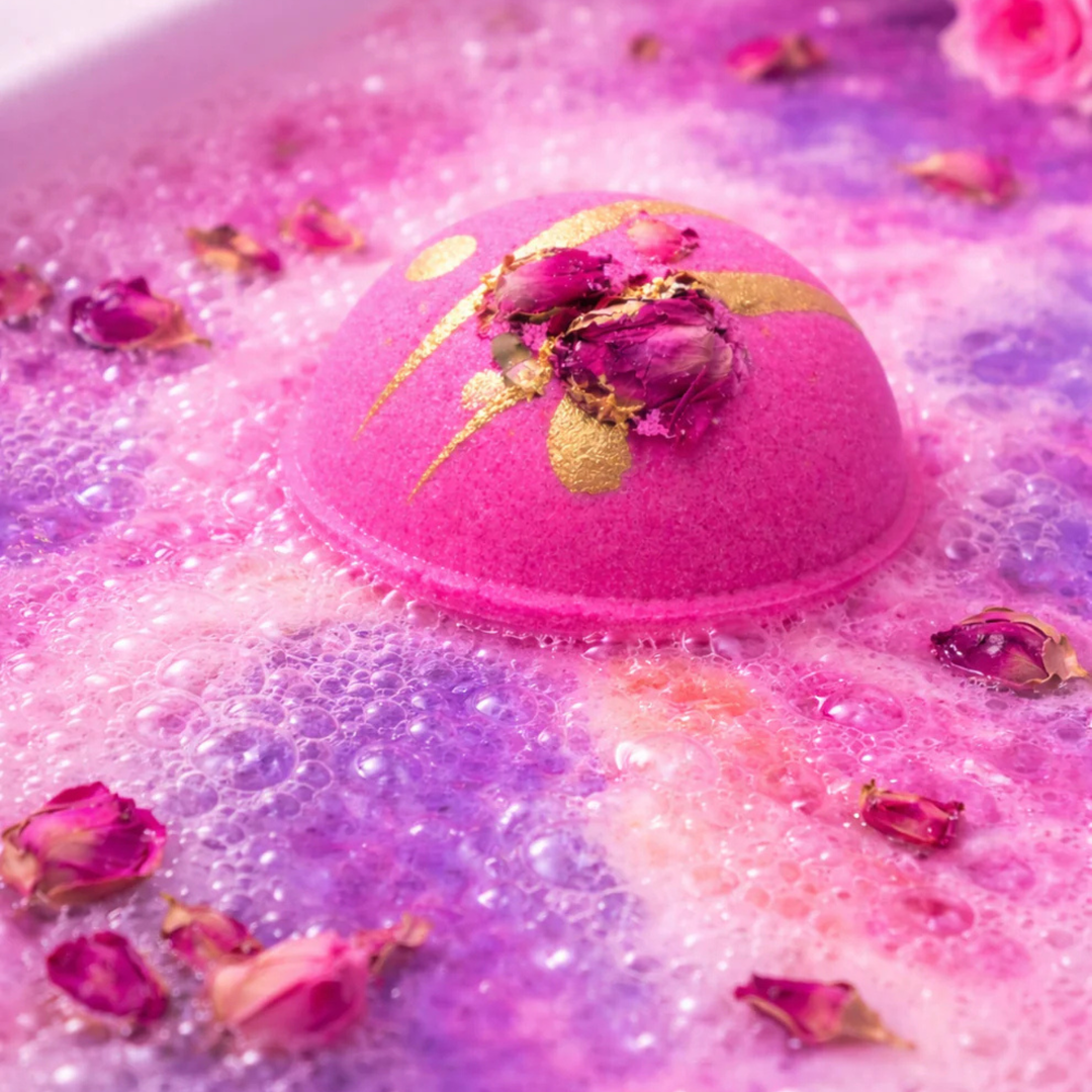 Pink bath bomb topped with gold glitter and rosebuds bubbling in a water bath.