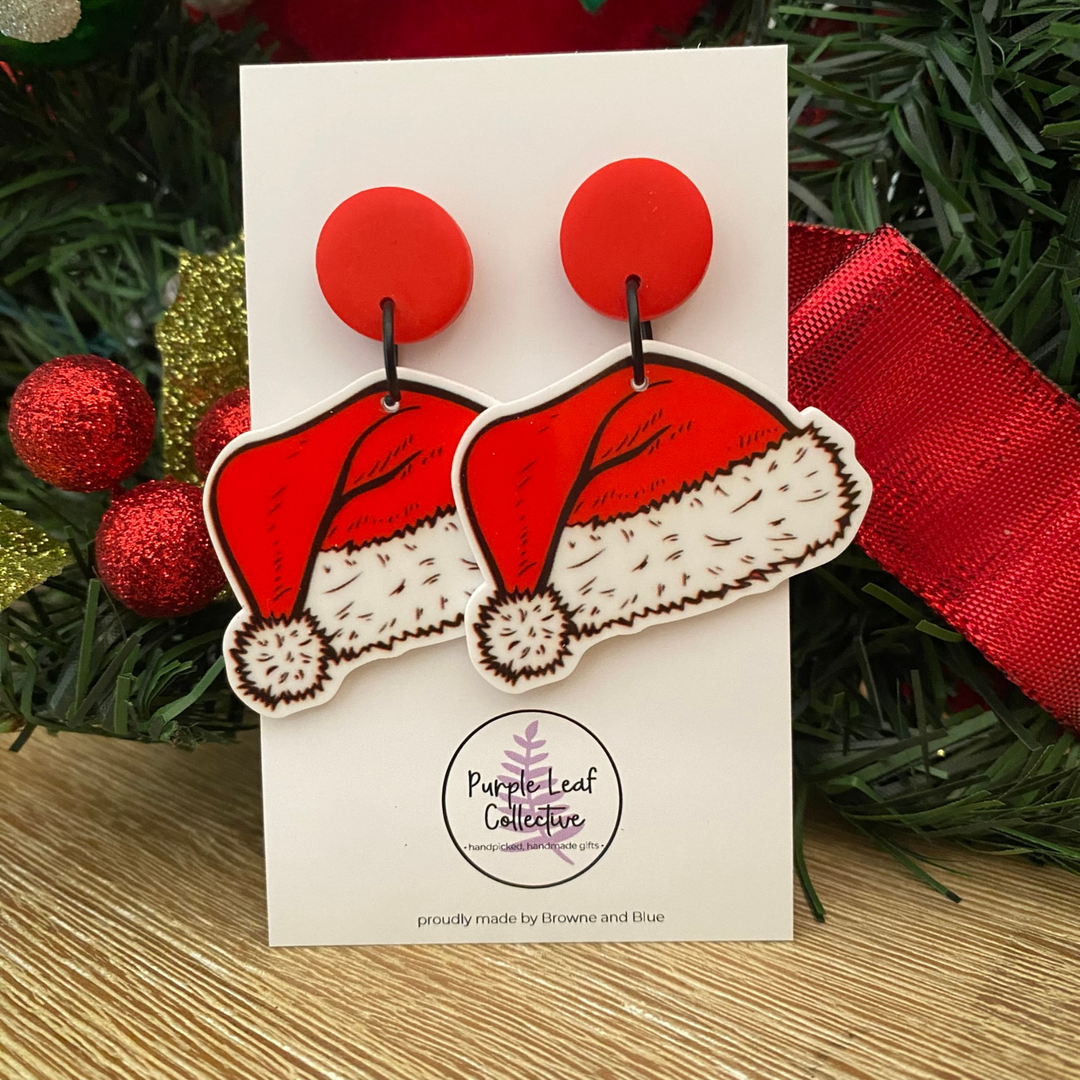 Red and white Santa hat earrings on a card with Christmas decorations in the background.