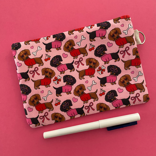 Reverse view of a zipper pouch with images of black and brown sausage dogs wearing jumpers and sunglasses on a pink fabric.