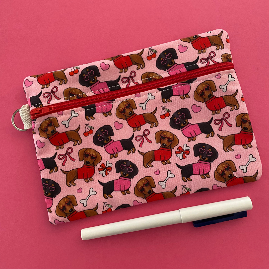 Front view of a zipper pouch with images of black and brown sausage dogs wearing jumpers and sunglasses on a pink fabric.