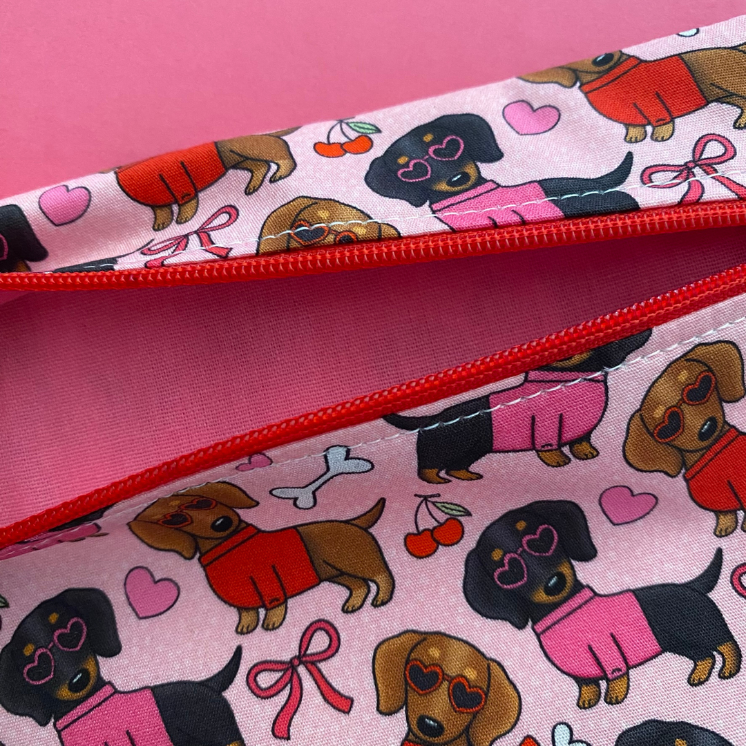 Zipper pouch with images of black and brown sausage dogs wearing jumpers and sunglasses on a pink fabric with vibrant pink linging.