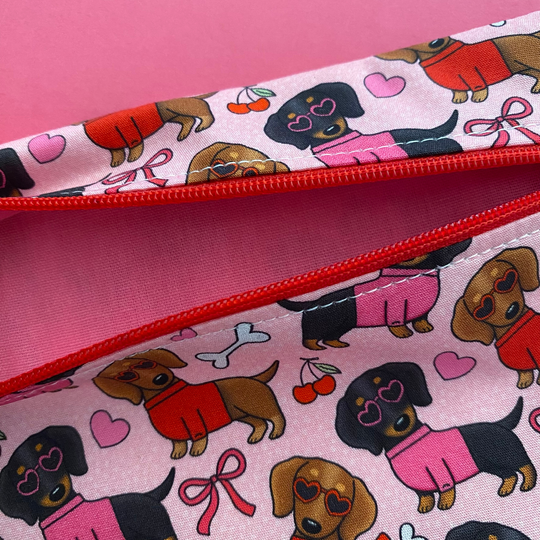 Zipper pouch with images of black and brown sausage dogs wearing jumpers and sunglasses on a pink fabric with vibrant pink linging.