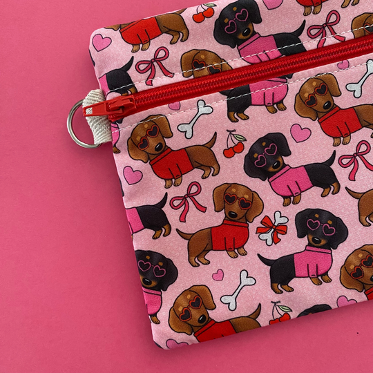 Zipper pouch with images of black and brown sausage dogs wearing jumpers and sunglasses on a pink fabric showing the D-ring.