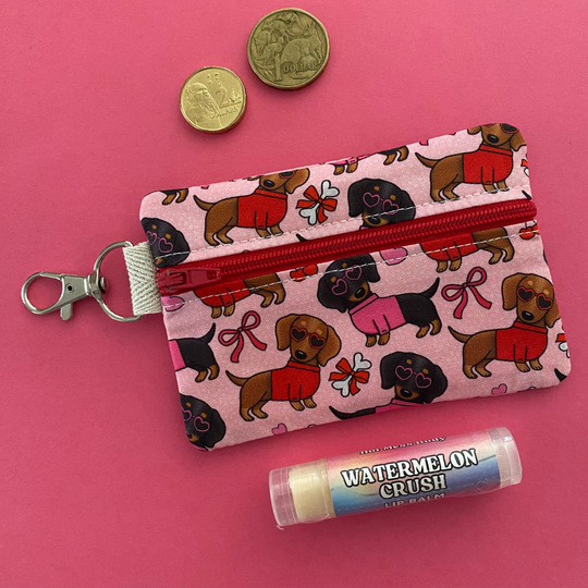 Front view of a mini pouch with images of black and brown sausage dogs wearing jumpers and sunglasses on a pink fabric.
