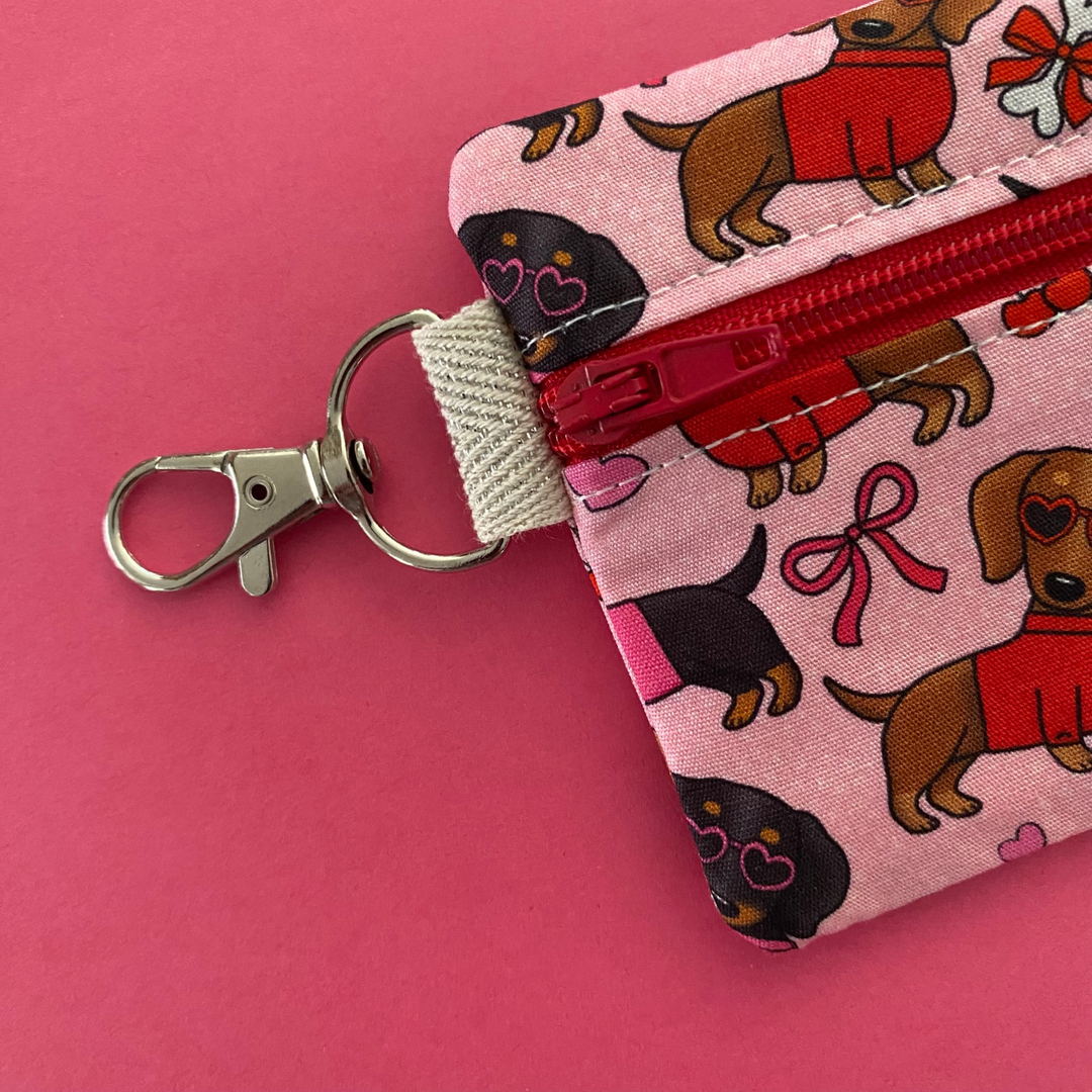 Mini pouch with images of black and brown sausage dogs wearing jumpers and sunglasses on a pink fabric showing the clasp.