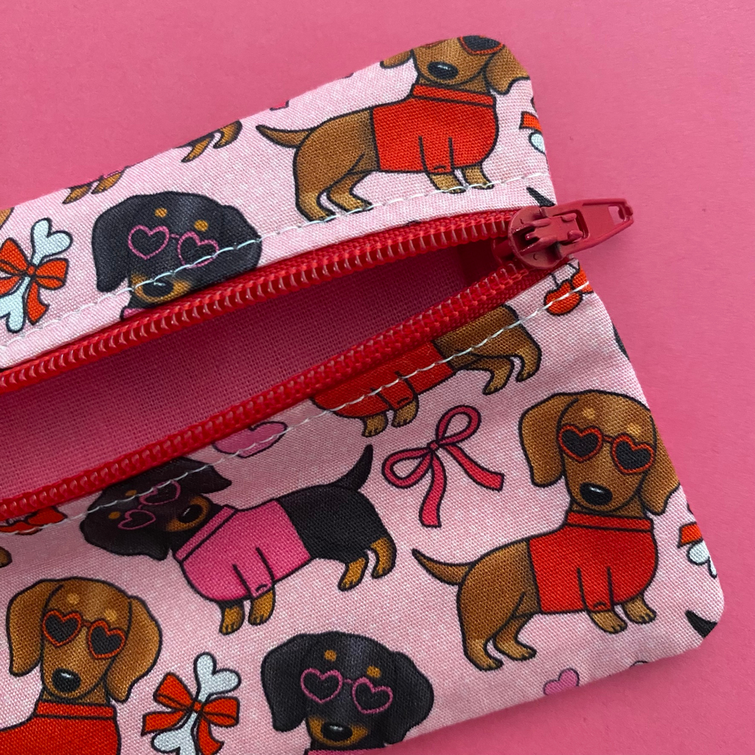 Mini pouch with images of black and brown sausage dogs wearing jumpers and sunglasses on a pink fabric showing the vibrant pink lining.