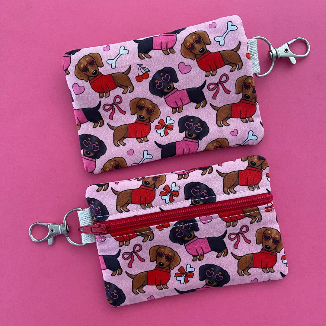 Reverse view of a mini pouch with images of black and brown sausage dogs wearing jumpers and sunglasses on a pink fabric.