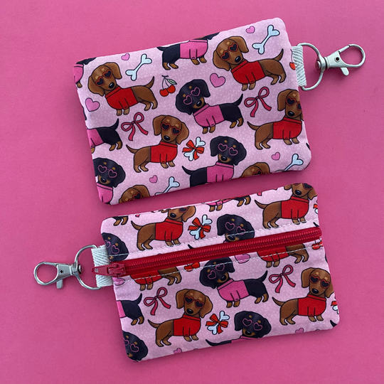 Reverse view of a mini pouch with images of black and brown sausage dogs wearing jumpers and sunglasses on a pink fabric.