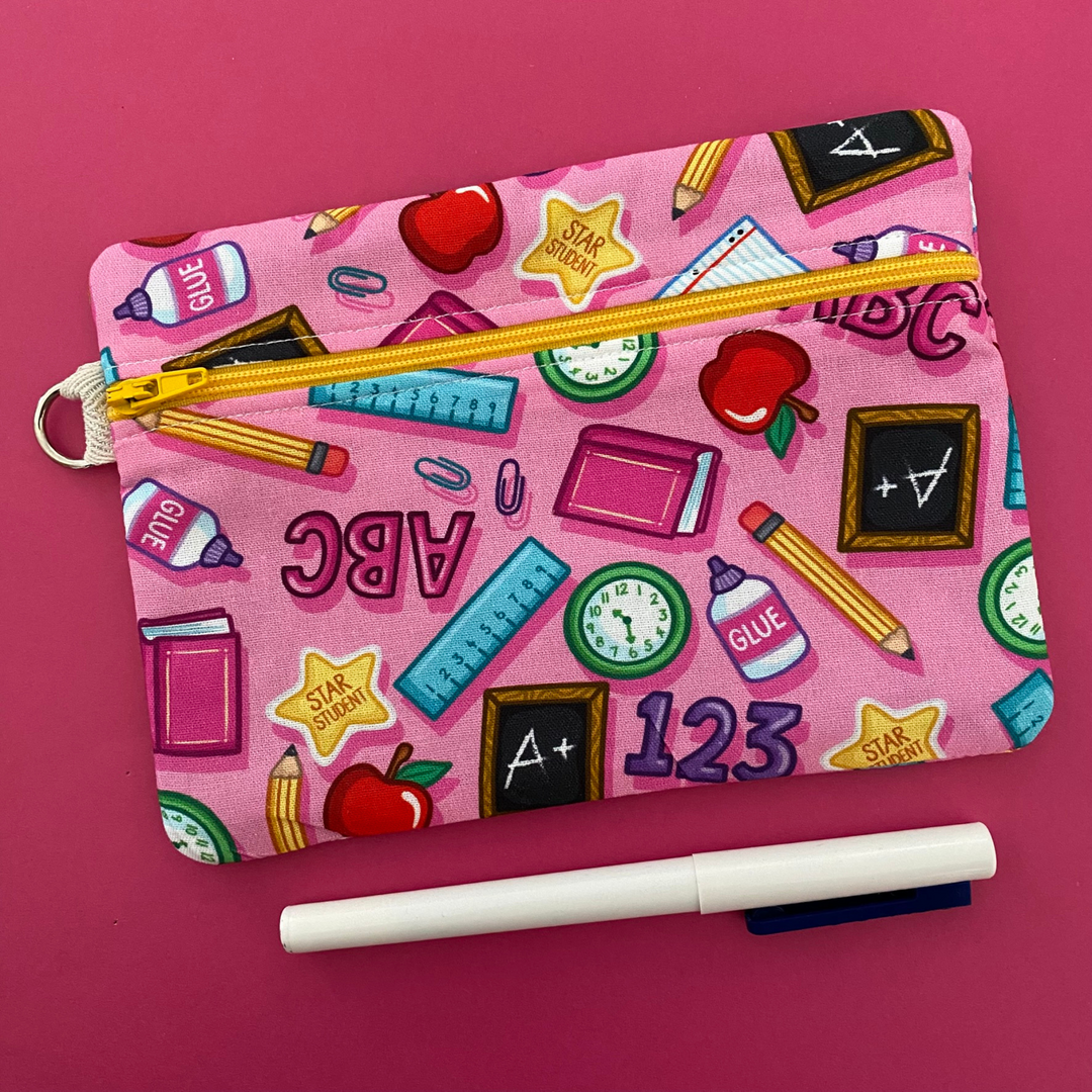 Zipper pouch adorned with playful illustrations of apples, rulers, pencils, glue, and books on pink fabric. Planner accessory for school or work. Perfect for small essentials.