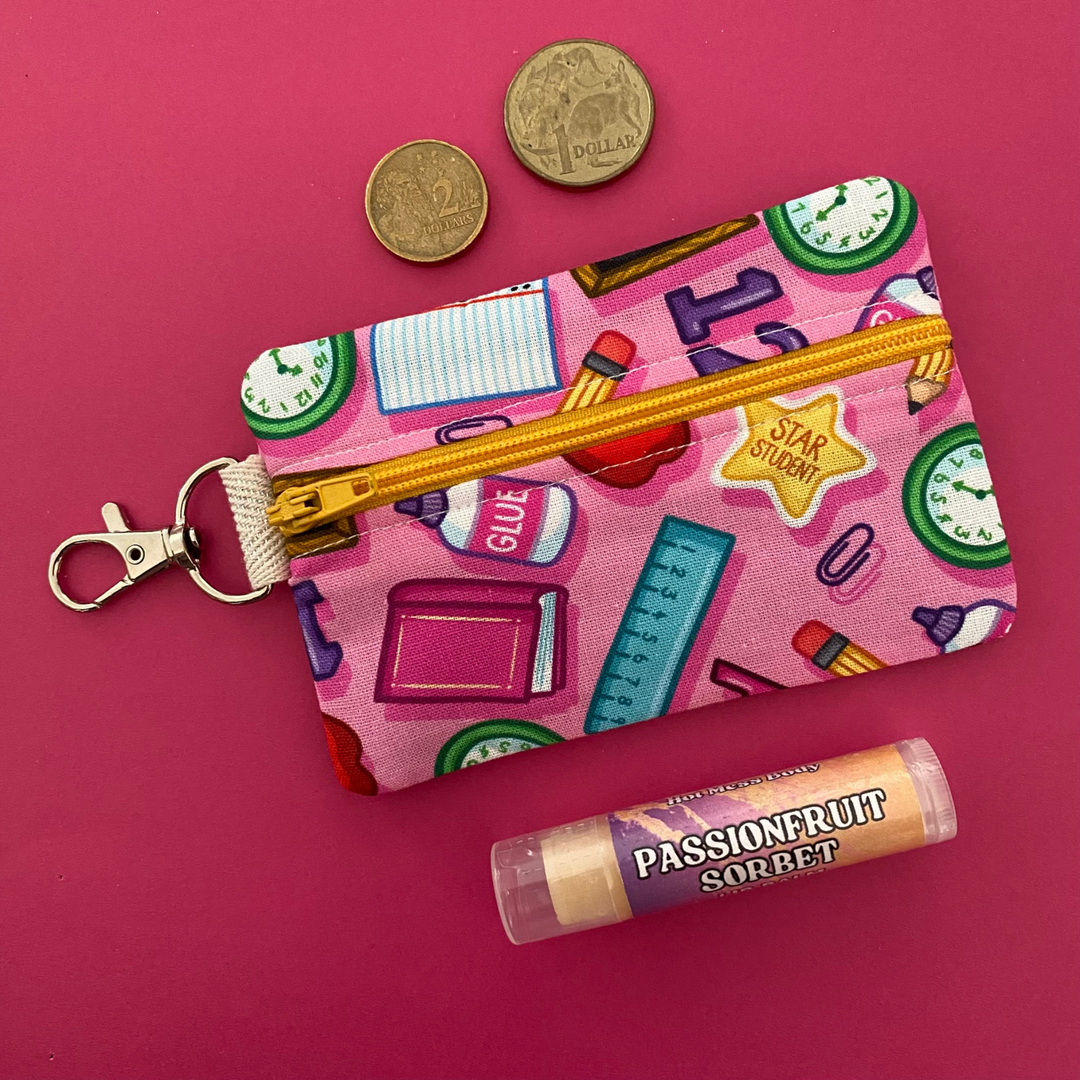 A small clip coin purse perfect for coins and small essentials featuring playful illustrations of apples, rulers, pencils, glue, and books especially loved by teachers, teacher aides, and early childhood educators.